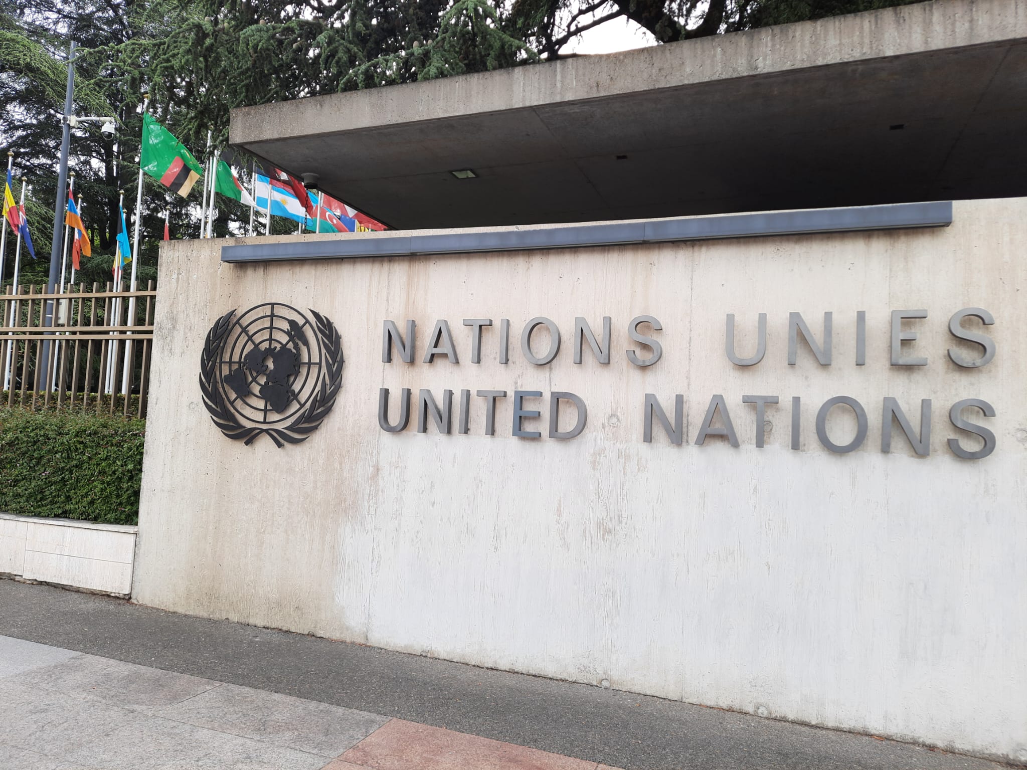 EN: Close-up of a concrete wall featuring the United Nations emblem and the inscription “Nations Unies – United Nations” at the entrance to the UN headquarters in Geneva, with member state flags in view.