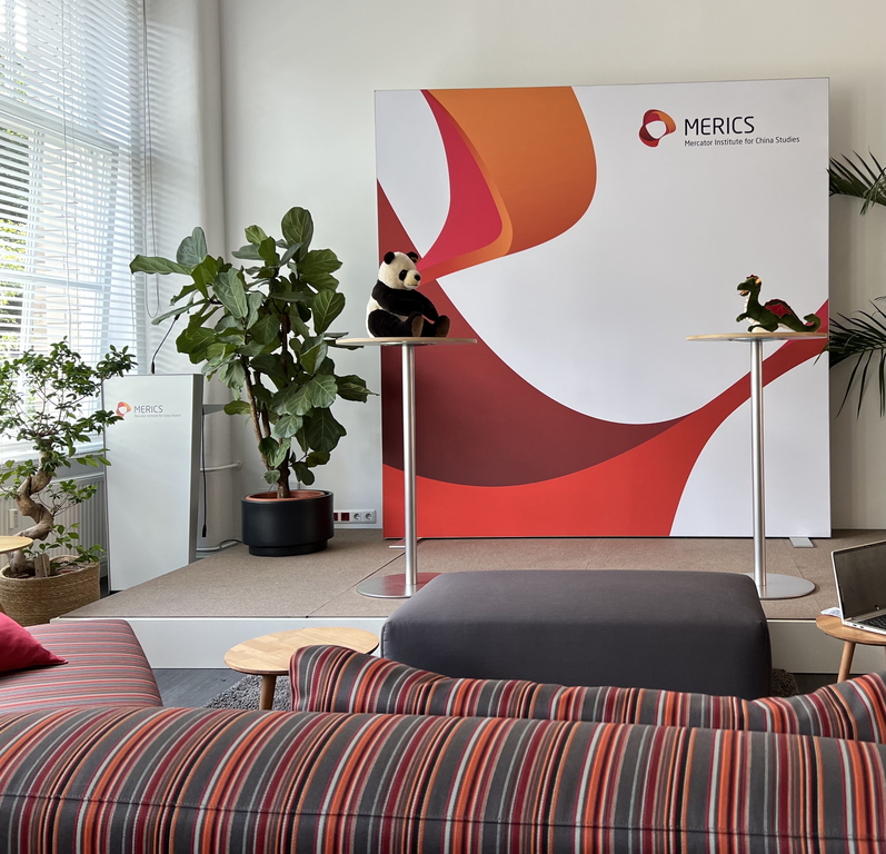 A wall with MERICS logos, in front of which are two tall cocktail tables. On one table lies a panda mascot, and on the other a dragon mascot.