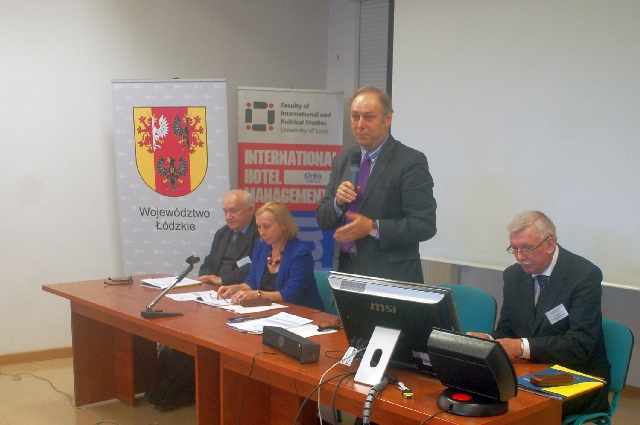 A panel table with three people sitting and one standing and speaking with a microphone
