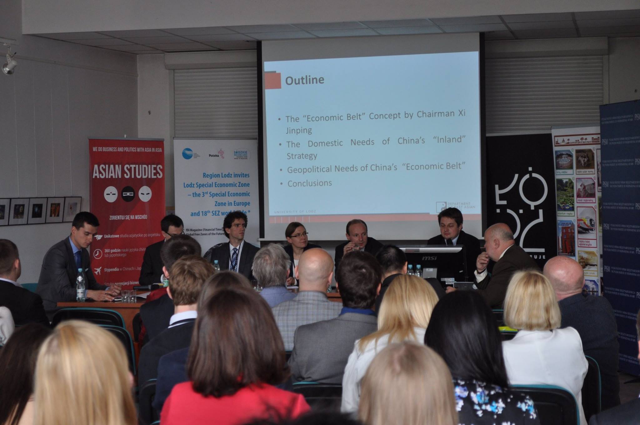 Panel discussion at a conference – speakers seated at a table with a presentation displayed on a screen behind them.