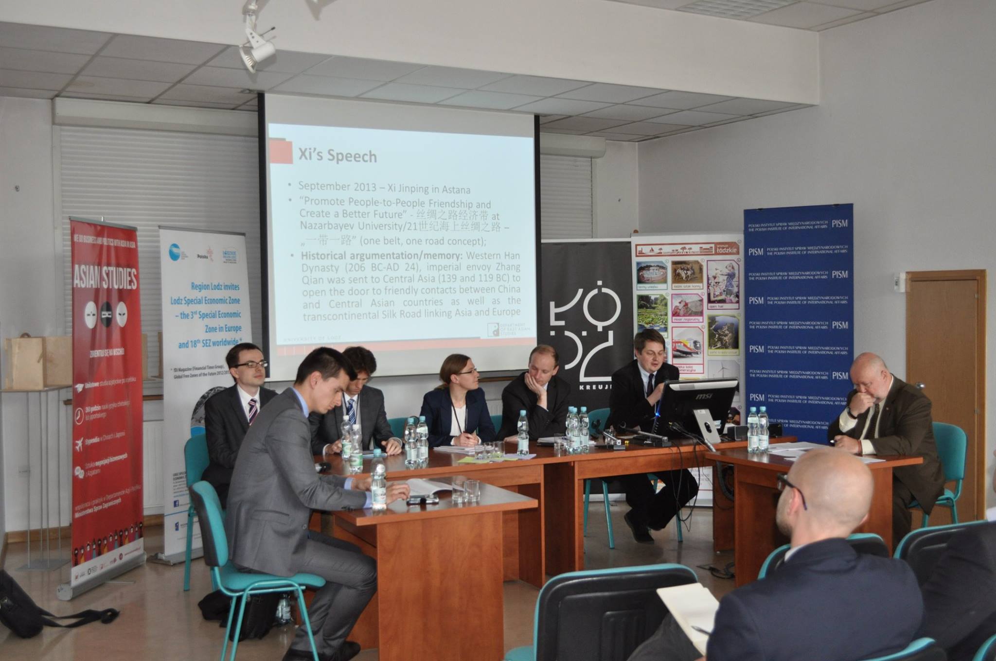 Panel discussion at a conference – speakers seated at a table with a presentation displayed on a screen behind them.