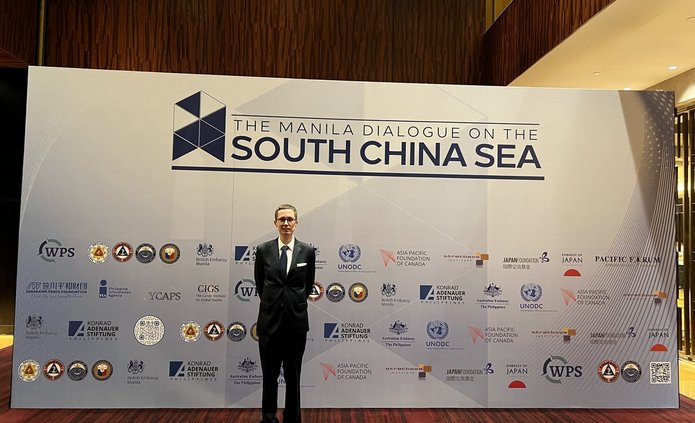 Mężczyzna w garniturze stoi przed ścianą z logotypami i napisem „The Manila Dialogue on the South China Sea” podczas konferencji w Manili.