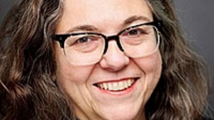 Studio headshot of a woman with long dark hair and glasses, wearing a dark blazer over a gray top, smiling in front of a neutral gray background.